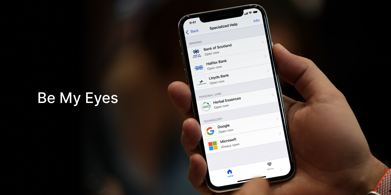 A hand-held iPhone with Specialized Help home screen displaying a list of companies providing Be My Eyes support, including Google, Herbal Essences, Lloyds Banking Group and Microsoft.